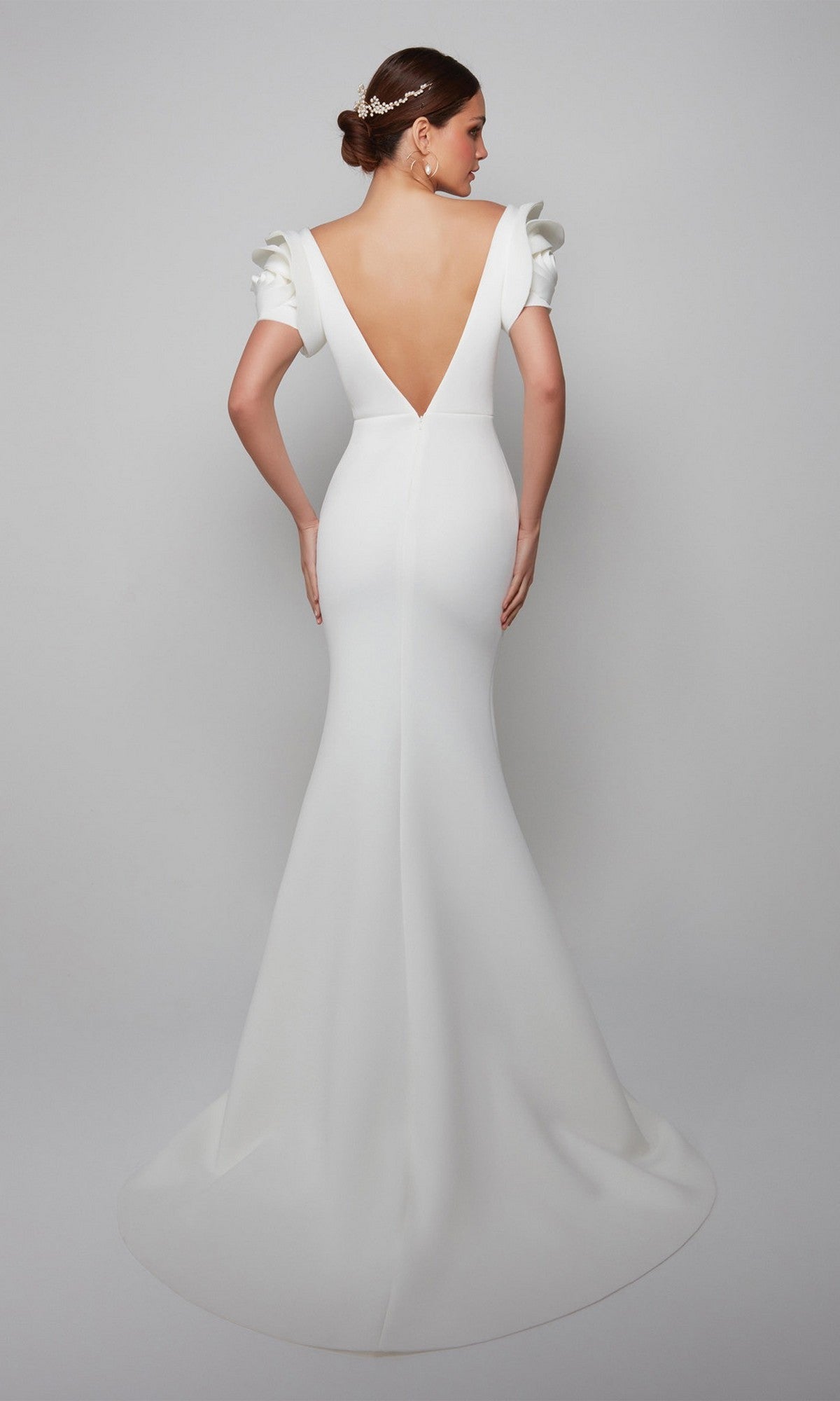 A woman in the Alyce 7066 long formal dress—white, fitted, with a mermaid silhouette, deep V-back, and short ruffled sleeves—stands facing away against a plain light background.